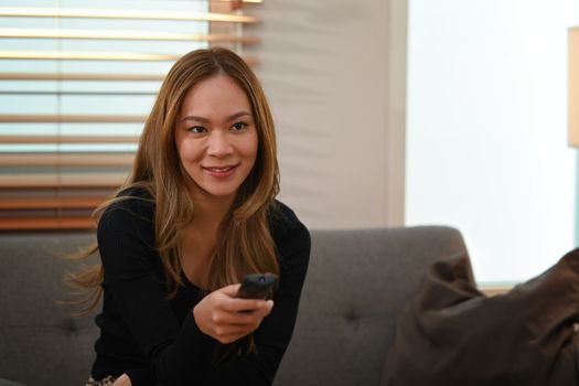 Pleased young woman with remote controller watching TV, spending leisure time on weekend at home
