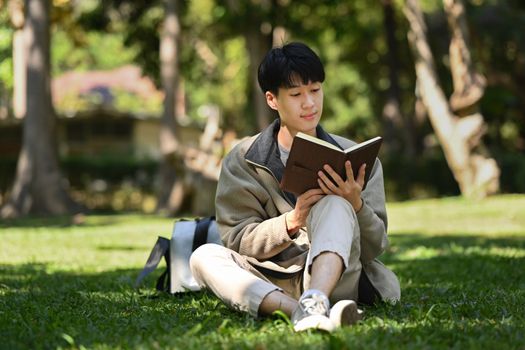 Happ young asian man reading interesting book while while sitting on green grass at sunny beautiful day