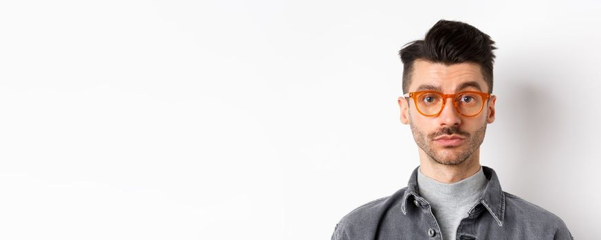 Attractive brunette man with moustache, wearing stylish glasses and look serious at camera, no emotion pokerface, standing against white background