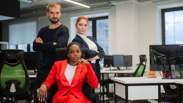 Caucasian red-haired woman, bearded Caucasian man stand behind a seated African American young woman in the office.
