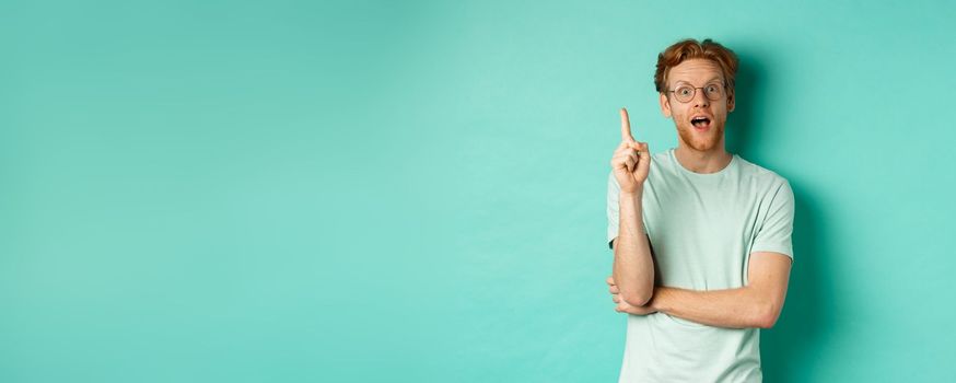 Excited young man with ginger hair in glasses, raising index finger, pitching an idea, standing over mint background