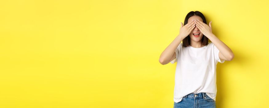 Happy asian woman waiting for surprise gift, close eyes and smiling, expecting something, standing over yellow background