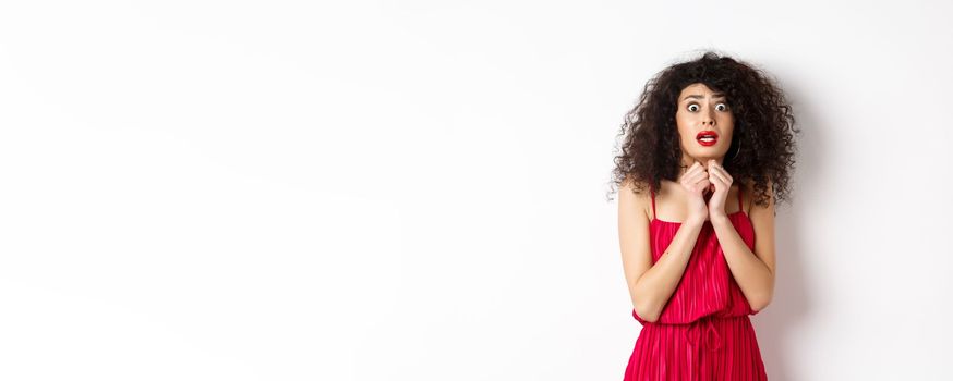 Scared caucasian woman trembling from fear, wearing red dress and staring anxious at camera, standing over white background