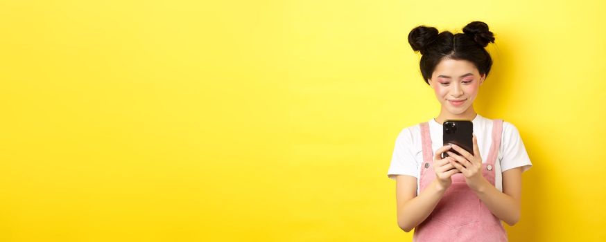 Stylish asian girl with glamour makeup using mobile phone, smiling at screen, standing on yellow background