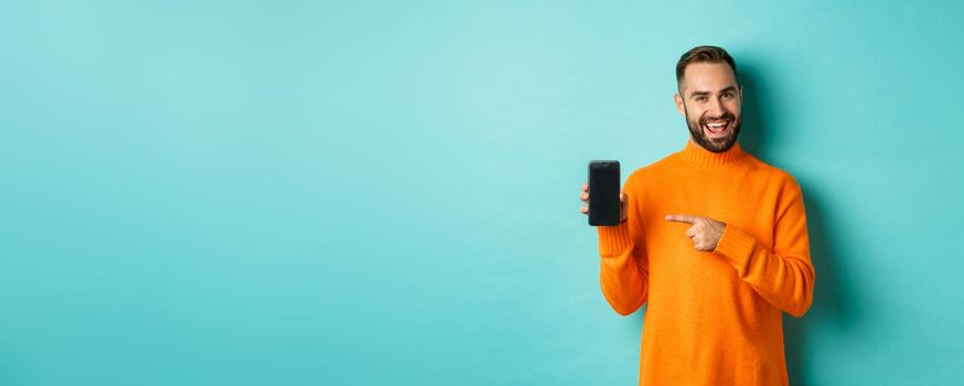 Handsome bearded man in orange sweater, pointing finger at mobile phone screen, showing application smartphone, standing over light blue background