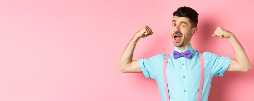 Strong and funny guy with french moustache, flexing biceps and winking at camera, show-off his strengths, standing over pink background