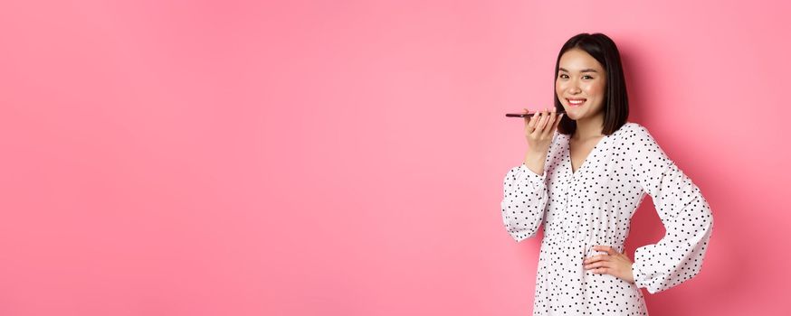 Beautiful korean woman talking on speakerphone, recording voice message and smiling happy, standing over pink background