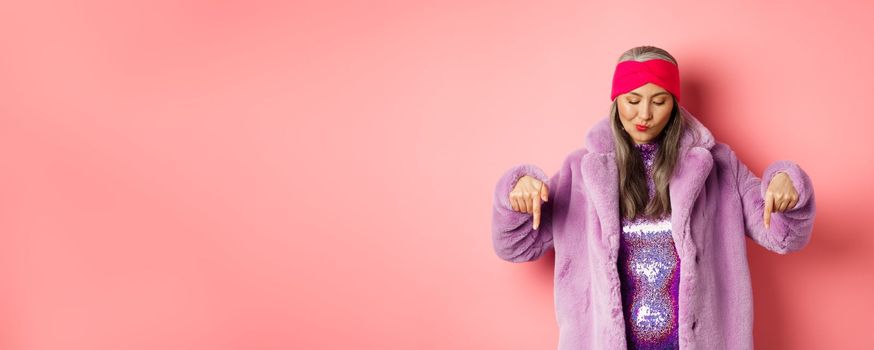 Fashion and shopping concept. Funky asian senior woman in trendy faux fur coat and disco dress, looking and pointing down, standing over pink background