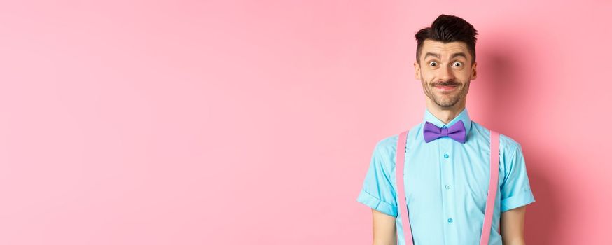 Smiling caucasian man looking excited, standing in classy bow-tie and shirt for romantic date, pink background