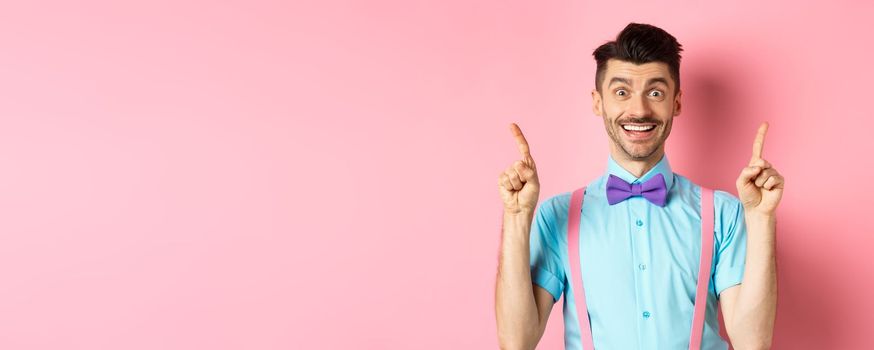 Portrait of cheerful young guy with moustache and bristle pointing fingers up, showing top logo, smiling happy at camera, standing on pink background