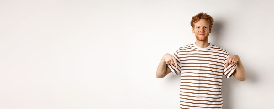 Handsome male model with ginger hair and bristle, pointing fingers down and smiling, showing advertisement, white background