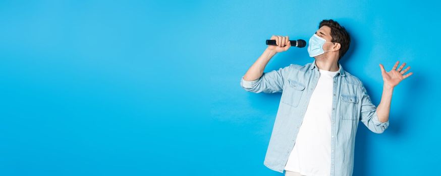 Handsome guy in medical mask singing in microphone, standing over blue background