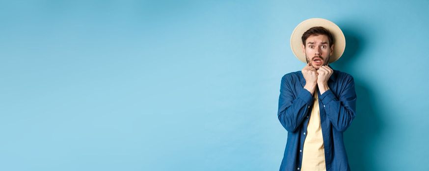 Scared young tourist trembling from fear, looking at something scary on summer vacation, wearing straw hat, standing on blue background