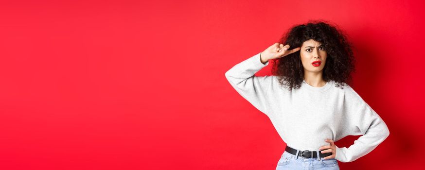 Are you stupid. Annoyed and confused girl with curly hair, pointing at head and scolding person being crazy or strange, standing on red background