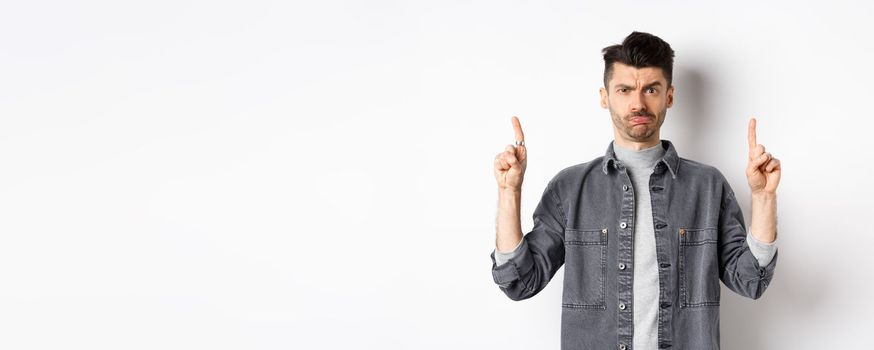 Confused funny guy with moustache pointing fingers up at something strange, frowning and pouting puzzled, standing on white background