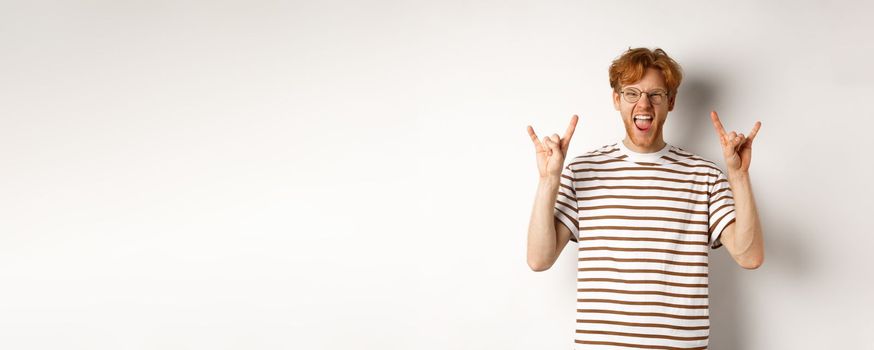 Funny and happy redhead man having fun, showing rock-n-roll horn and sticking tongue, enjoying party, standing over white background