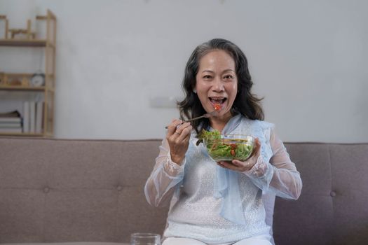 Portrait, Cheerful Asian 60s aged woman having breakfast in her living room, eating healthy salad vegetables mix bowl.