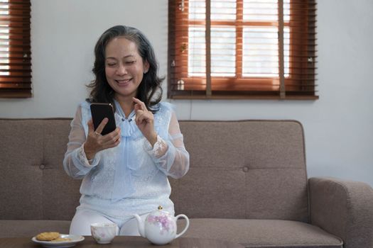 Cheerful 60s retired Asian woman using her mobile phone to chat with her grandchild while relaxing in her living room.