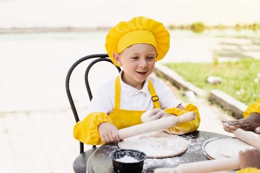 Handsome cook child in yellow chefs hat and apron yellow uniform holding dough roller and cooking dough outdoor.