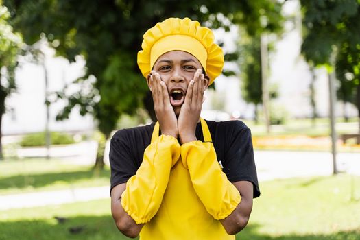 Shocked black african teenager cook in chefs hat and yellow apron uniform hold cheeks and surprise. Creative advertising for cafe or restaurant.