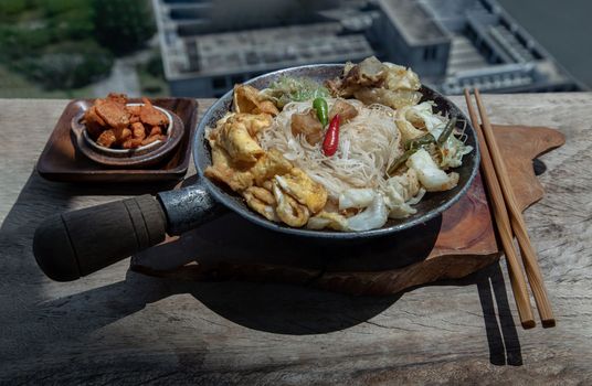 Stir fried rice vermicelli with pork dumplings, egg and vegetable in small steaming iron pot serve with crispy pork crackling or pork scratching.