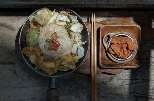 Stir fried rice vermicelli with pork dumplings, egg and vegetable in small steaming iron pot serve with crispy pork crackling or pork scratching.