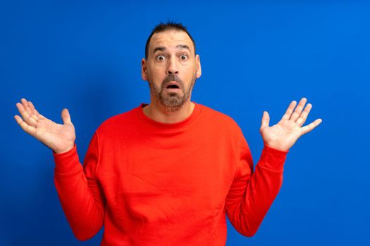 Amazed and excited latin man looking at camera with a shocked expression feeling shocked from advertising shopping promotion, amazing betting win standing isolated on a blue background