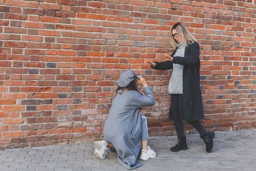 Girl takes picture of her female friend in front of brick wall in urban street copy space - photographer and youth urban lifestyle concept