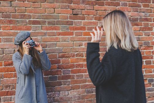 Girl takes picture of her female friend in front of brick wall in urban street - photographer and youth urban lifestyle concept