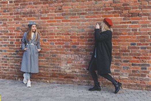 Girl takes picture of her female friend in front of brick wall in urban street - photographer and youth urban lifestyle concept