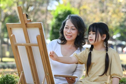 Happy middle aged grandma painting picture with lovely little grandchild, enjoying leisure weekend activity together outdoor
