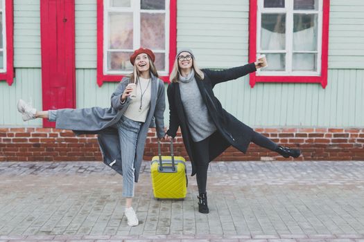 Two cheerful funny tourist women smiling and fool around and with suitcases on city street in autumn or spring time - travel and vacation concept