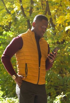 Millennial african american male walking on city street autumn park holding mobile phone for blogging. Smiling man student strolling on urban setting with cellular - social network concept