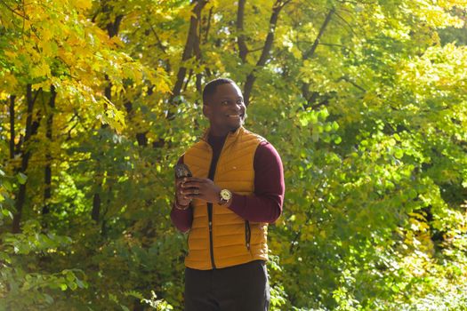 Millennial african american male walking on city street autumn park holding mobile phone for blogging copy space. Smiling man student strolling on urban setting with cellular - social network concept