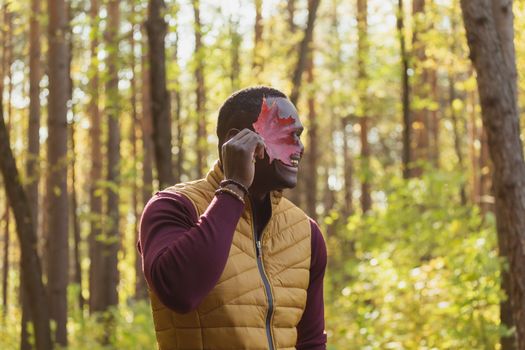 Portrait of african american man covering his face with autumn maple leaf. Autumn nature. Seasonal fall fashion.