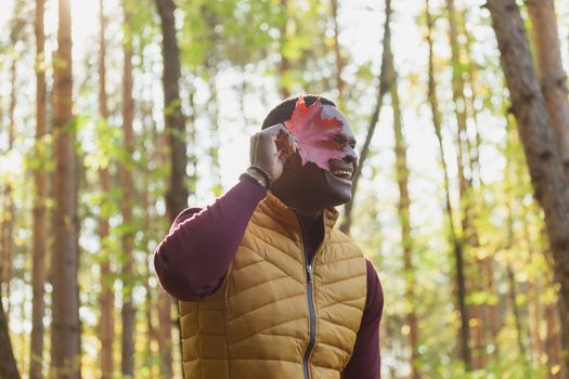 Portrait of african american man covering his face with autumn maple leaf. Autumn nature. Seasonal fall fashion.