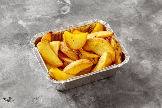 Aluminium foil container with baked potato wedges on gray stone tabletop