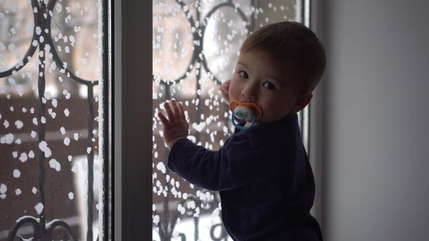 The child looks out the window and knocks on it. A one-year-old son stands at the window in winter.