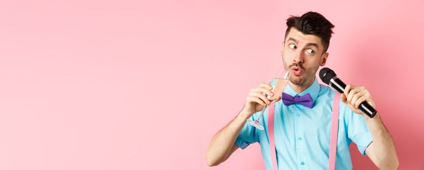 Party and festive events concept. Funny guy singing karaoke, performing song with microphone and drinking champagne from glass, standing on pink background
