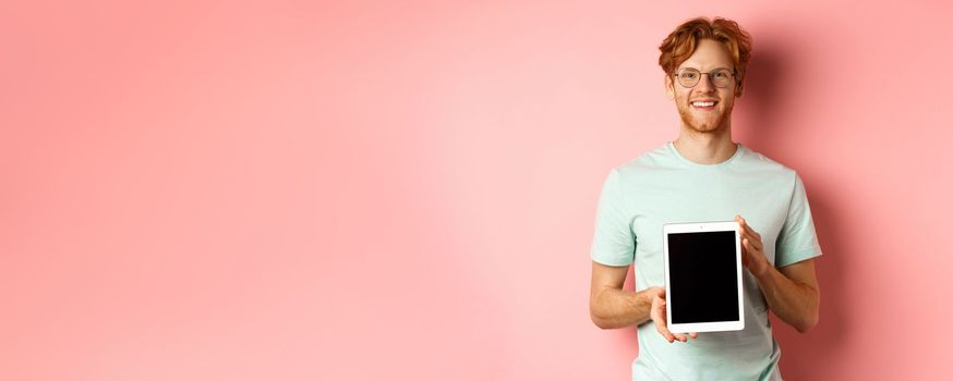 Funny redhead guy in glasses demonstrate digital tablet screen, smiling at camera, standing over pink background