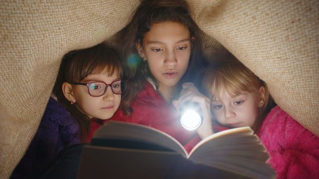 Children girl kids under blanket reading interesting fairytale story book using flashlight at home