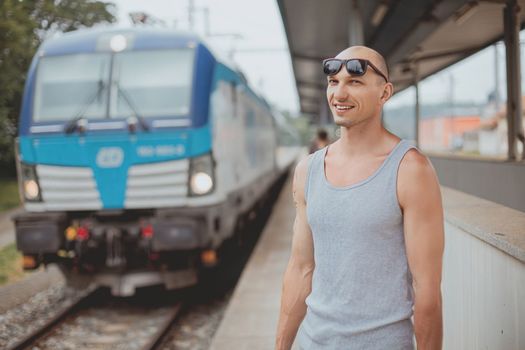 Young man in a t-shirt on the platform waiting for a train using mobile phone. Man by train station platform suburban with bag and mobile phone. Traveling by train.