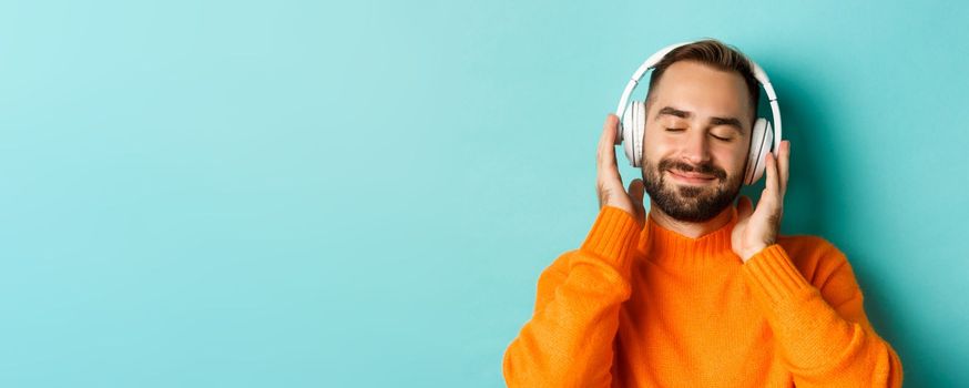 Close-up of handsome modern man listening music in headphones, standing in orange sweater over turquoise background
