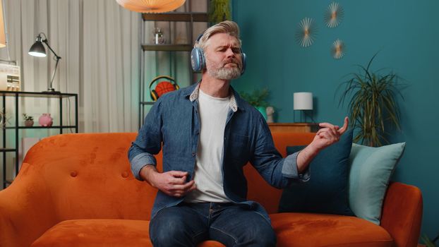 Adult man in headphones dancing on sofa at home listening favorite music playing on imaginary guitar