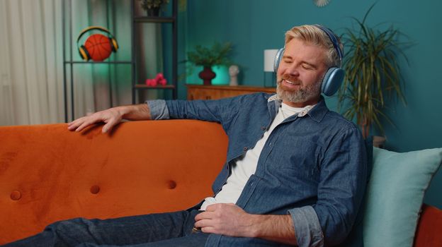 Happy mature man in wireless headphones relaxing sitting on sofa at home listening favorite music