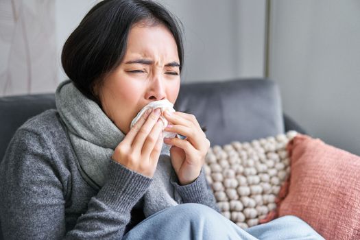 Close up of young korean woman staying at home with cold, sneezing in napking, has runny nose, concept of illness, health and influenza