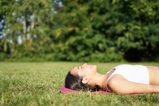 Young fitness girl lying on sport mat on lawn, breathing and meditating in park in sportswear