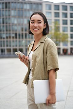 Vertical shot of young asian digital nomad, girl talks on mobile phone and walks on street with laptop. Young woman remote worker going to coworking space