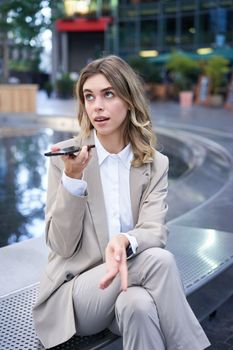 Smiling business woman record voice message, speaking into microphone on mobile phone, sitting near fountain on street