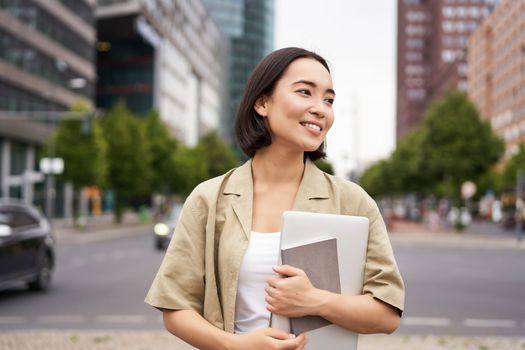 Portrait of beautiful asian woman standing on street with laptop and notebook, going to university or work, commute route